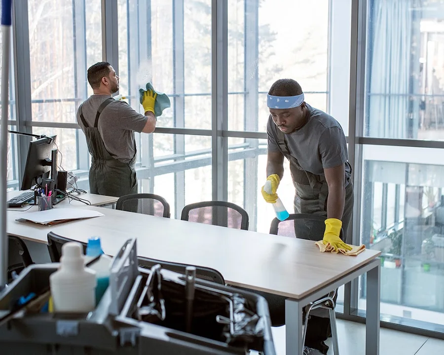 Zwei Reinigungskräfte bei der Unterhaltsreinigung in einem Büro: Schreibtische und Fensterfront